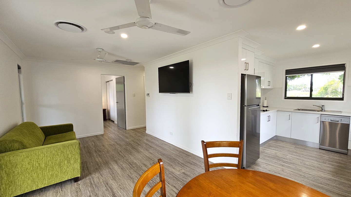 To the left is a door leading to the bedroom. Adjacent, a green couch rests against the middle wall, with a flatscreen TV mounted on the wall. To the right, a kitchen area is equipped with kitchen equipment, drawers, cabinets, and a sink. A round wooden table sits nearby. A window is positioned above the sink area. The floor is timber, the walls are white, and the ceiling features ceiling fans and LED lights.