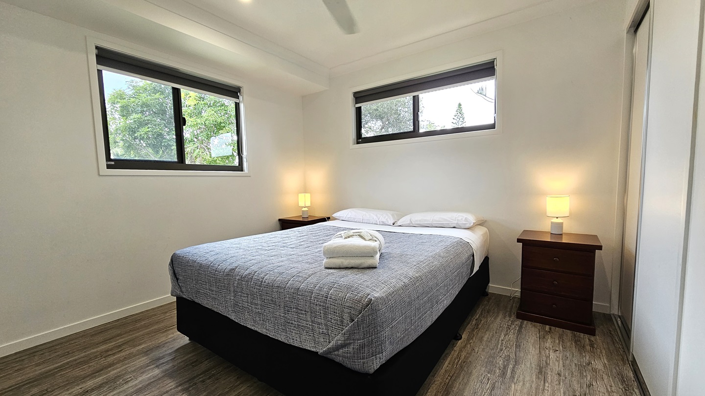 In the centre stands a white queen-size bed adorned with a grey cover. White pillows and a neatly folded white towel rest atop. Flanking the bed are two bedside tables, each with a lampshade. A window is situated on the central and left walls. The walls and ceiling are painted white, illuminated by LED lights and a ceiling fan. The floor is timber.