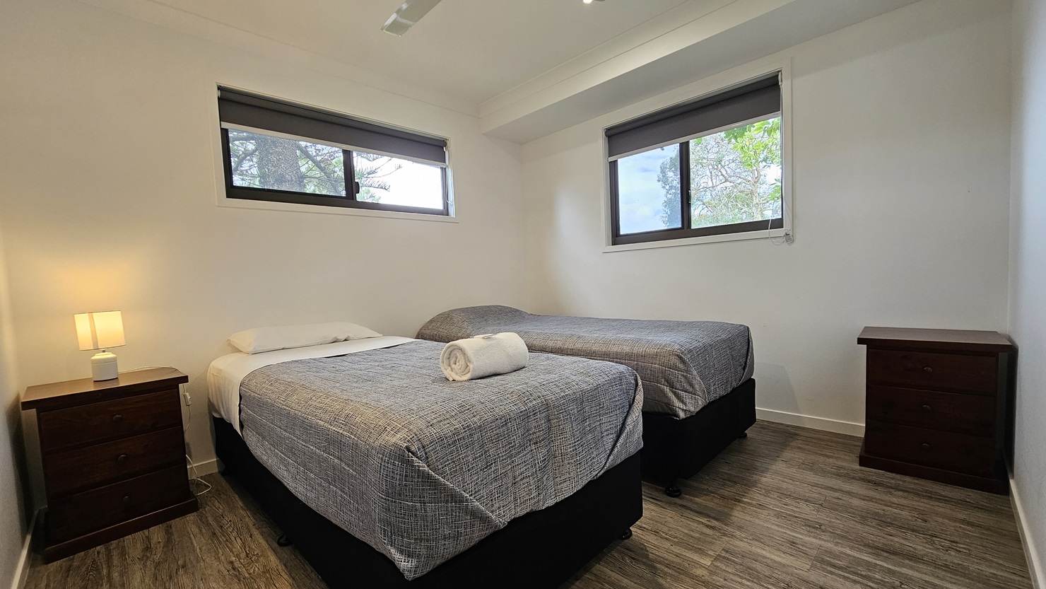 In the room, two single beds with grey covers stand opposite each other, each adorned with white pillows and a neatly rolled white towel. Windows are positioned on the middle and right walls. A bedside table with a lampshade is placed on the left corner, and another table on the right. The walls and ceiling are painted white, illuminated by LED lights and a ceiling fan. The floor is timber.