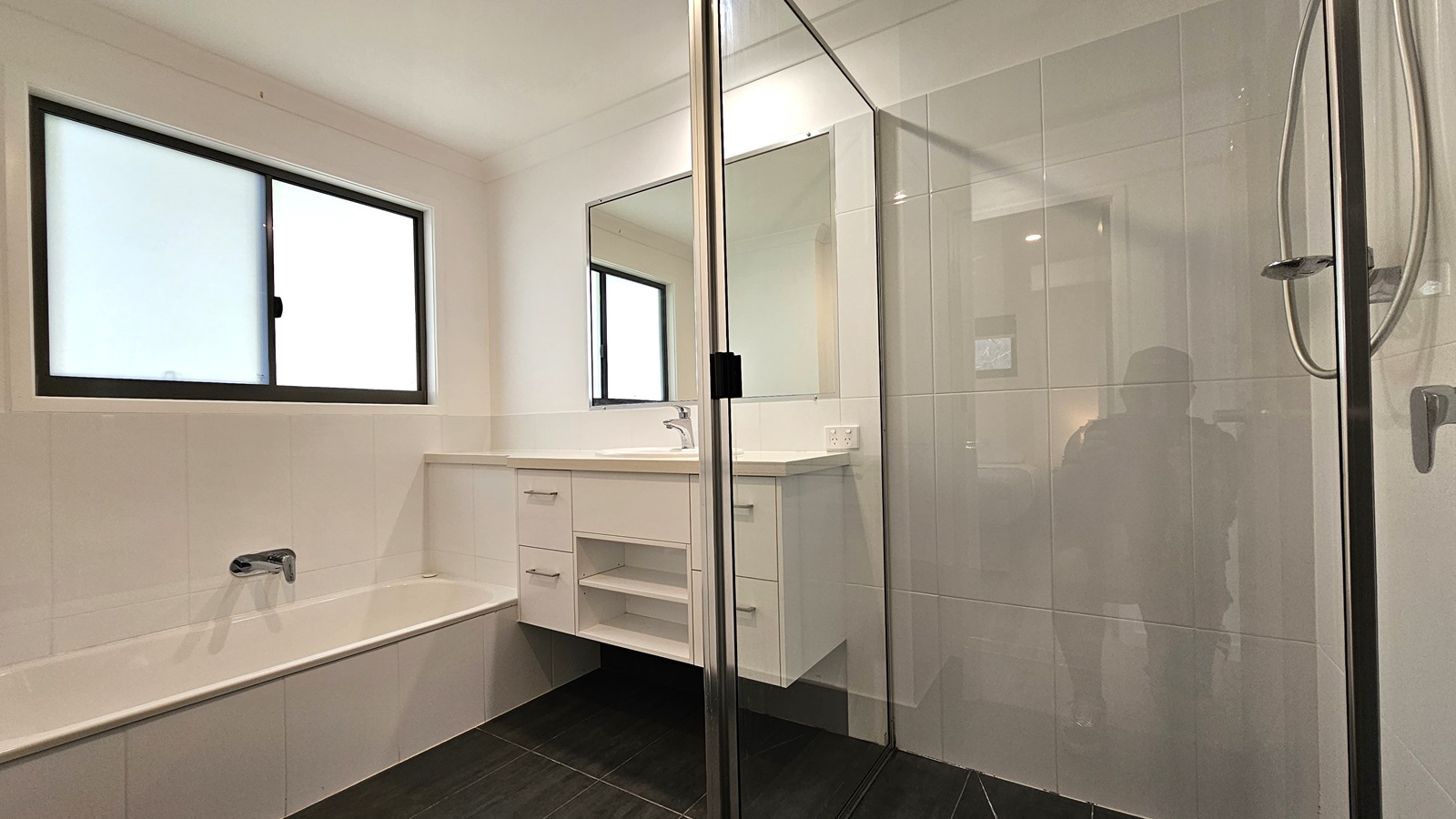 In the accessible bathroom, a shower with a handrail is positioned on the right, enclosed by a glass partition. Adjacent is a lavatory equipped with drawers and a mirror. On the left is a bathtub set against the wall, with a window positioned above it. The floor features grey tiles, while the walls and ceiling are white with LED lights.