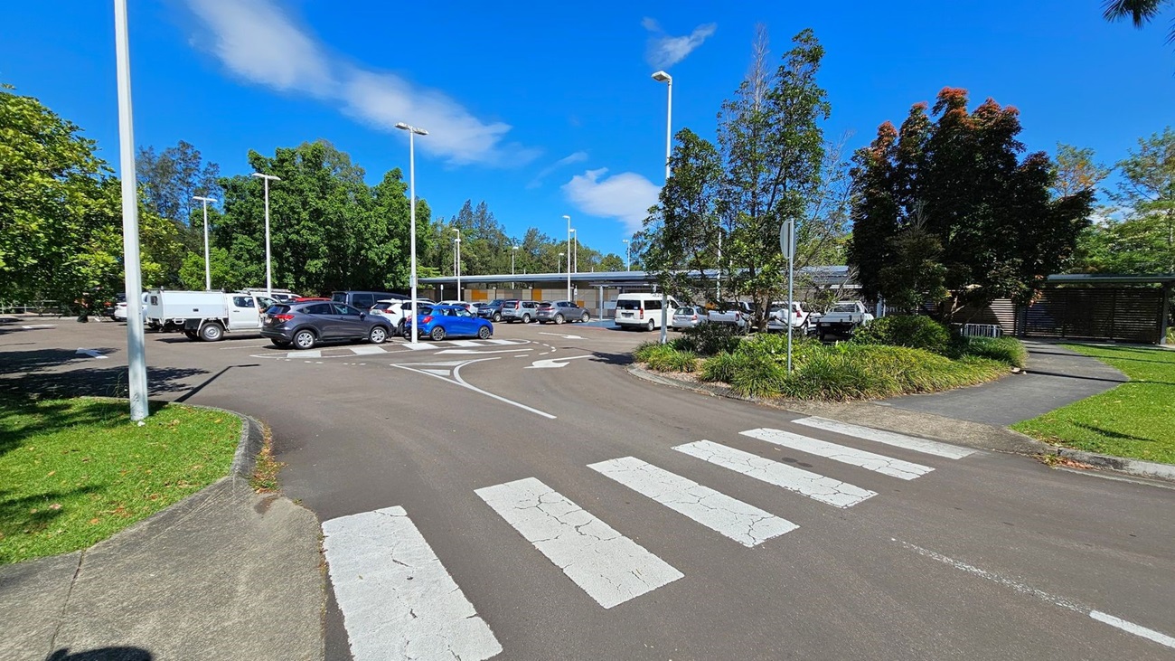 A car park with several vehicles, including a white utility vehicle and a blue car, is in the middle. The foreground has a zebra crossing on an asphalt road. On the left, there is a grassy area with a tree and a tall streetlight. On the right, there are more trees, a footpath, and a grassy patch. In the background, there is a low building with a covered walkway and more parked cars. Tall streetlights are scattered throughout the car park.