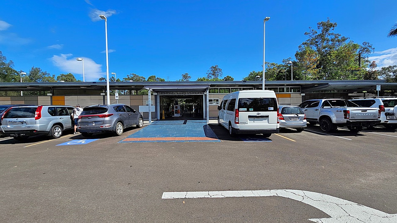 A car park with several vehicles, including a silver SUV, a grey SUV, a white van, and a white pickup truck. The car park has designated disabled parking spaces marked in blue. In the centre, there is a covered entrance to a building with a blue-tiled walkway leading to it. The building has a modern design with a mix of grey and beige panels. Trees and greenery are visible in the background. The ground is asphalt, and there are tall streetlights on either side.