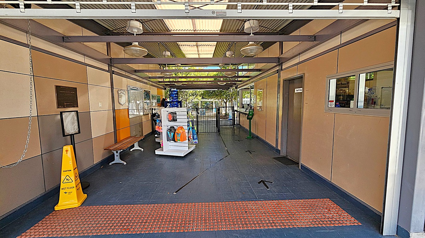 A corridor with a dark tiled floor and a ceiling with exposed beams and hanging lights. On the left, there is a yellow caution sign, a bench, and a wall with plaques. In the middle, a white display stand holds various items, and a gate is visible further back. On the right, there is a "Staff Only" door and a window with a counter. The background shows trees and outdoor light coming through the gate.