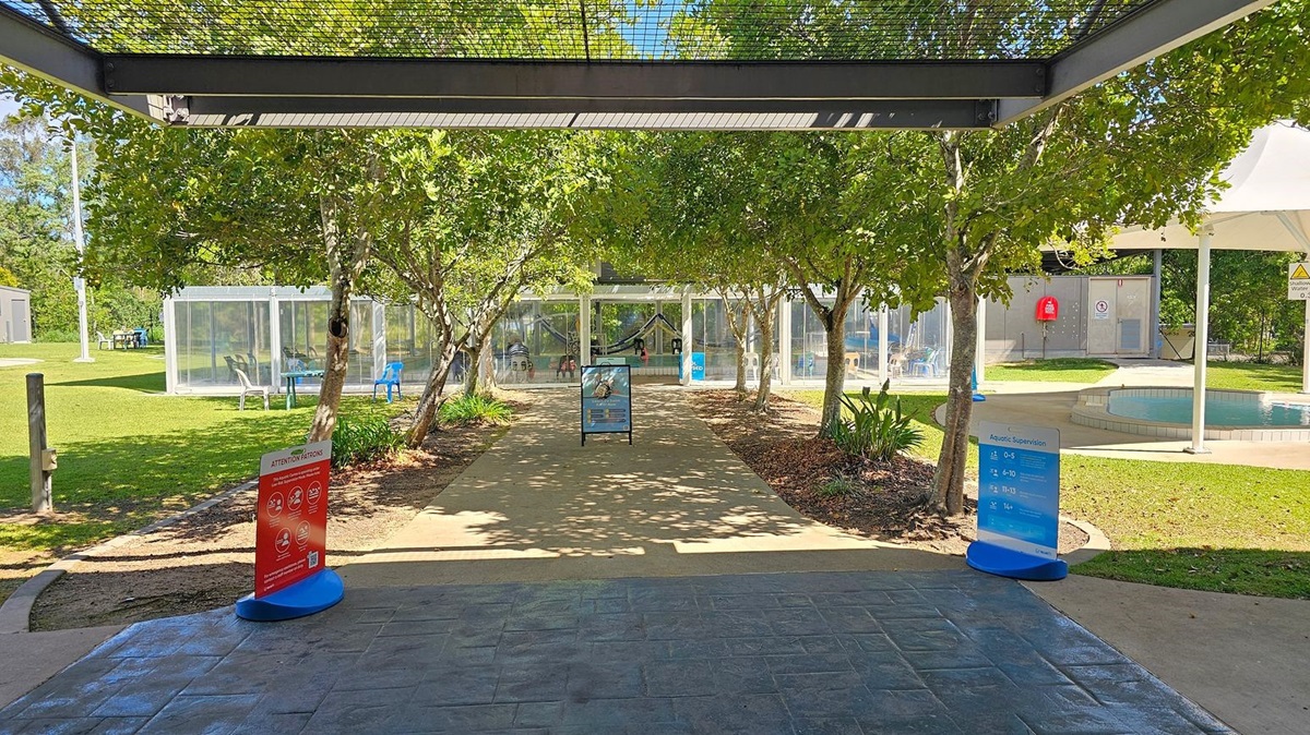 A paved pathway leads through a garden area with trees on both sides. In the foreground, there are two signs, one red and one blue, providing information. To the left, there is a grassy area with a transparent structure containing chairs. To the right, there is a small circular pool with a white canopy overhead. In the background, a glass building with people inside is visible. The floor is paved, and the ceiling is a metal grid structure.