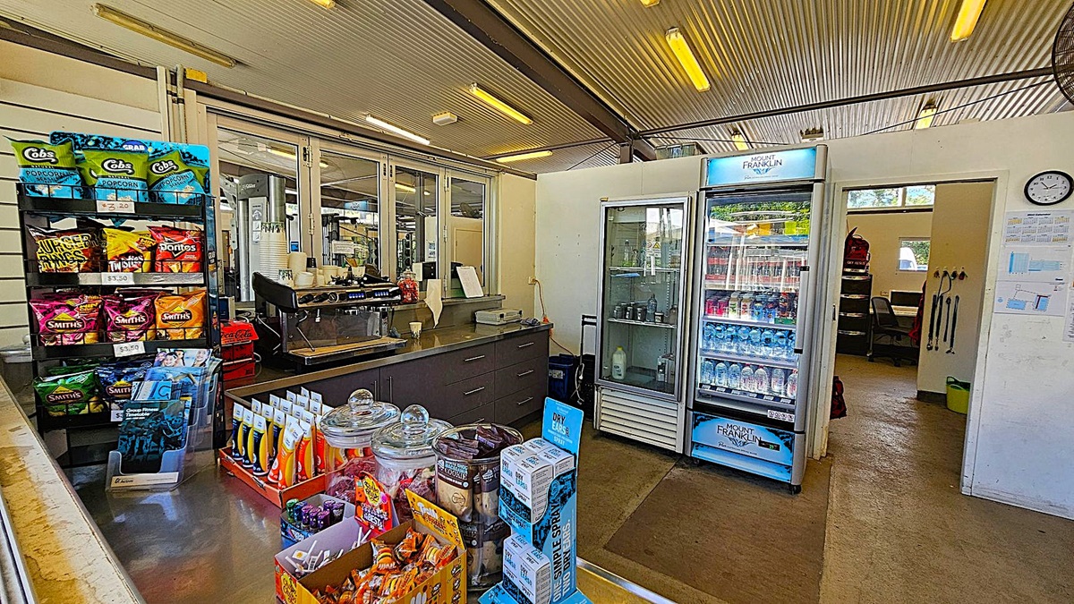A small shop interior with a corrugated metal ceiling and concrete floor. On the left, there is a display rack with various snack packets, including popcorn and crisps. In the middle, there is a counter with a coffee machine, jars of sweets, and other small items. On the right, there is a refrigerator stocked with bottled water and other drinks. Behind the refrigerator, there is an open doorway leading to a small office area with a desk and chair.