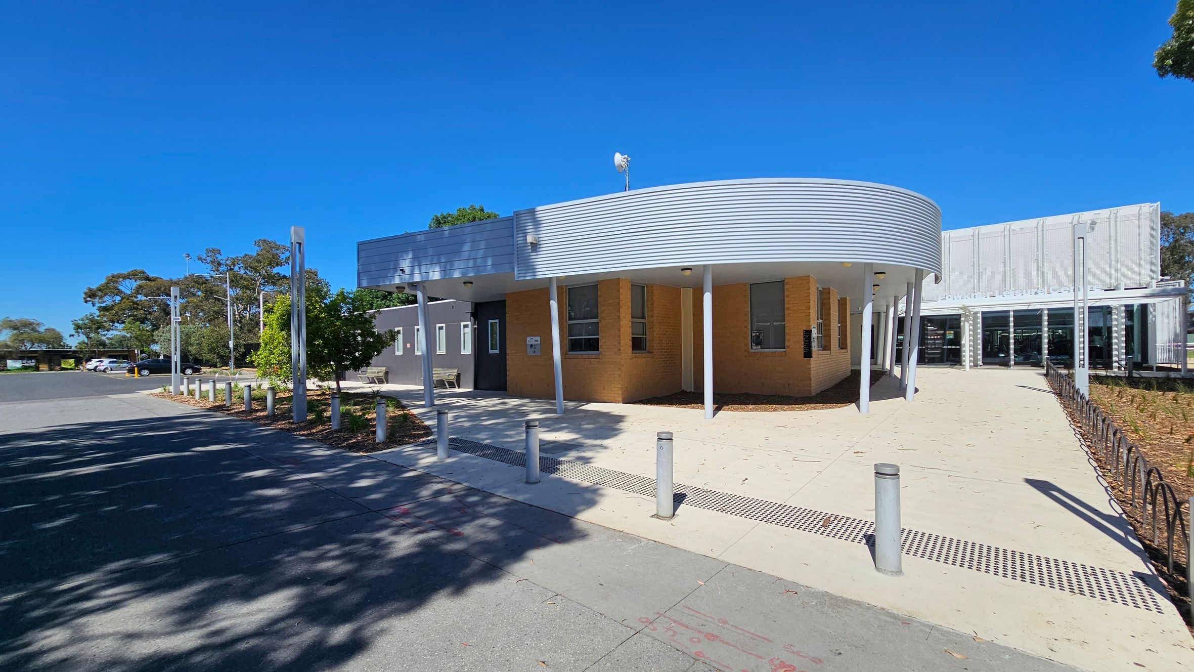 A street scene with a modern building on the left, featuring a curved entrance and large windows. In the centre, there's a playground with colourful equipment, surrounded by trees. On the right, several cars are parked along the street, including a red car in the foreground. The ground is a mix of pavement and mulch, with a bike rack near the building. Tall, slender light poles are positioned near the playground and building.