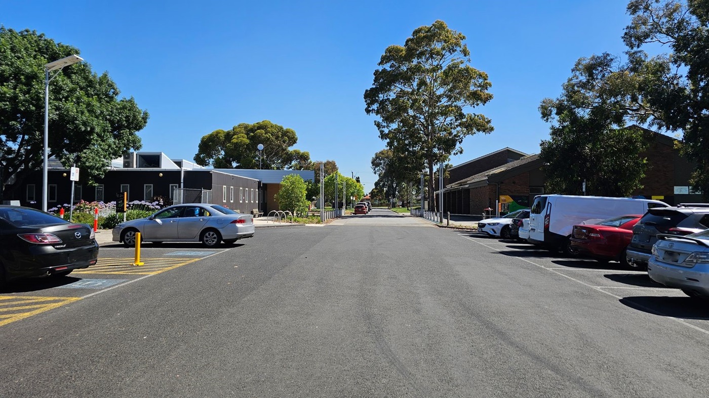 A wide asphalt road flanked by parked cars on both sides. On the left, a black building with a sloped roof is partially obscured by a large tree. On the right, a row of cars is parked next to a long, dark building. In the centre, the road extends into the distance, lined with trees and streetlights. The sky is clear and blue.