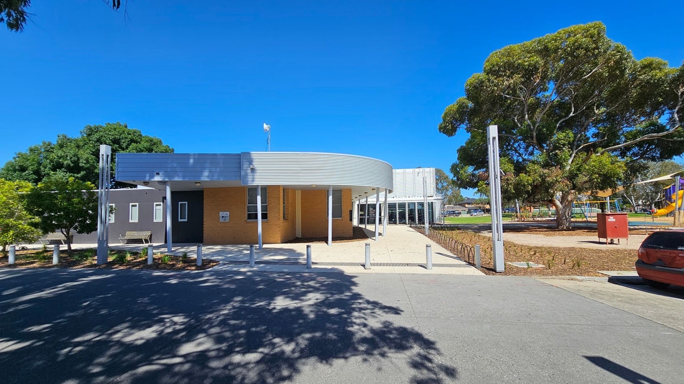 A modern building with a curved metal roof and brick walls is in the centre. To the left, there are trees and a bench. On the right, a large tree and a playground with slides and swings are visible. The foreground shows a paved area with shadows from the trees. A red car is parked on the right side.