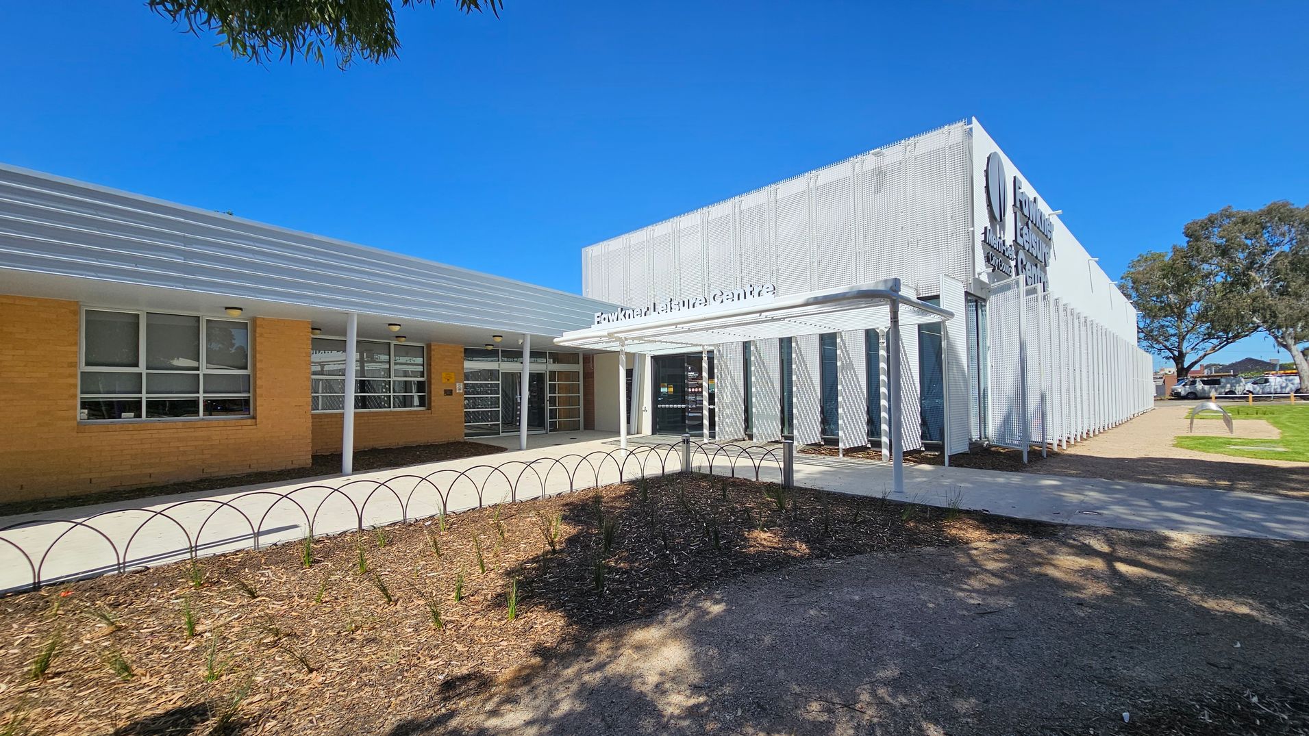 A modern building with a combination of brick and metal cladding. On the left, there are large windows set in a brick wall. The entrance is in the centre, featuring a white metal awning with the name "Fawkner Leisure Centre" above it. To the right, the building has a perforated metal facade. In the foreground, there is a landscaped area with mulch and small plants, and a curved metal bike rack. Trees and a car park are visible in the background.