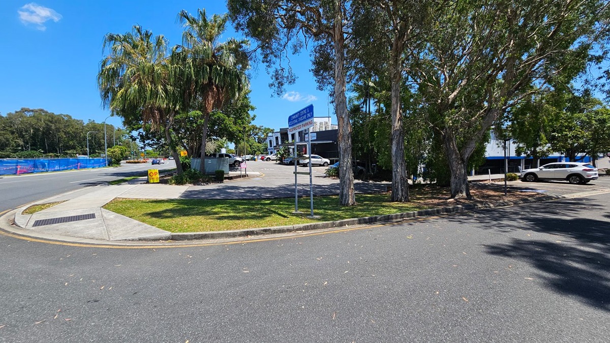 A street scene with a curved road in the foreground. On the left, there's a footpath and a grassy area with a sign that reads "OP SHOP." Palm trees and other greenery are present. In the centre, a parking area with several cars is visible, along with a blue sign indicating restricted parking. On the right, more trees provide shade, and a white car is parked. A building with a sign for a community centre and medical centre is in the background.