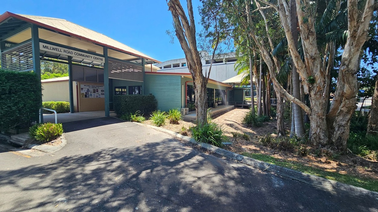 Millwell Road Community Centre with a sloped roof and a sign above the entrance. The building is light blue with a covered walkway. On the right, there are tall trees and a garden area with shrubs. The foreground shows a paved path with shadows from the trees.