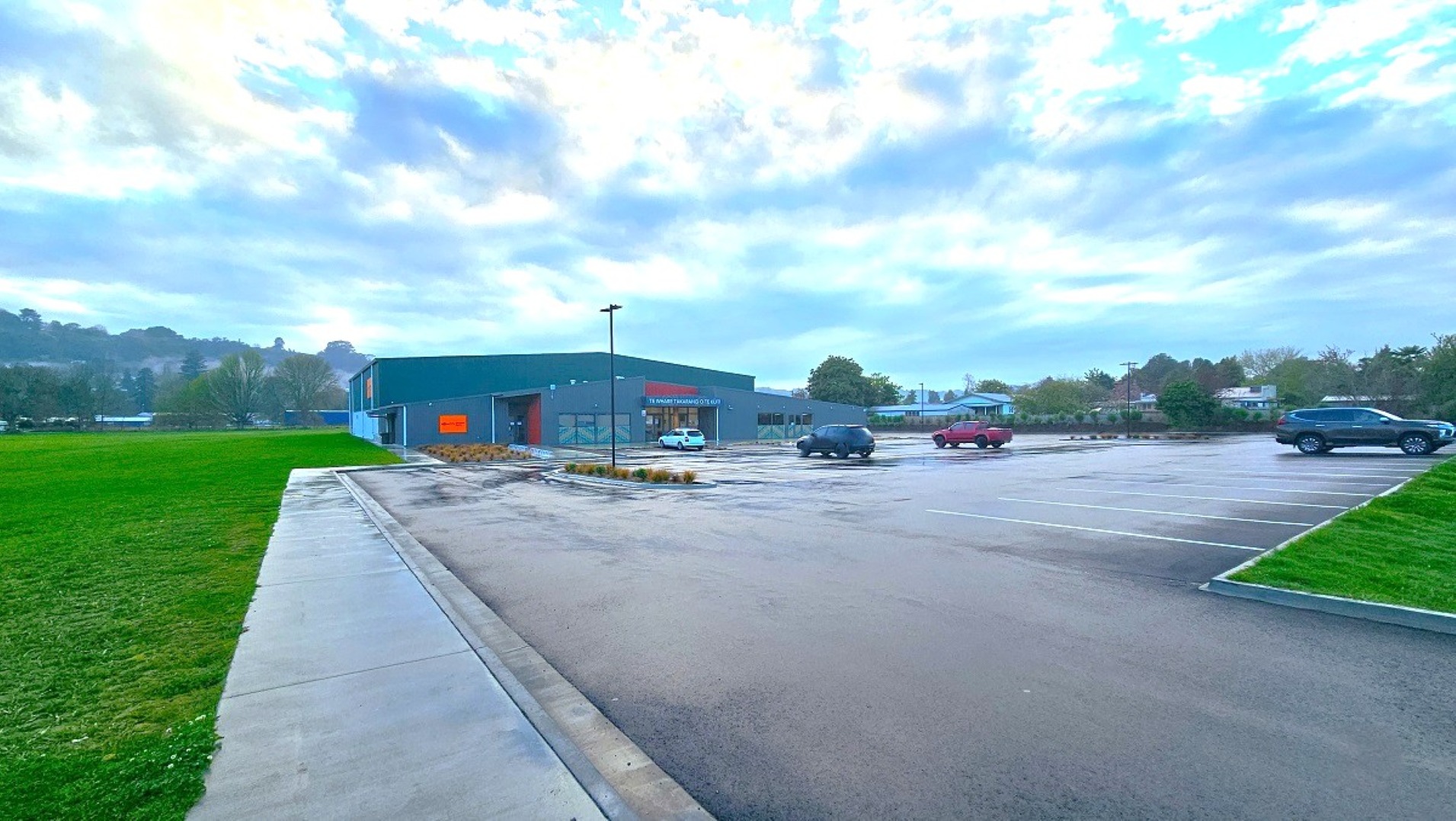 A large parking area with a few cars scattered around. On the left, there's a wide expanse of green grass. In the centre, a single-storey building with a dark green exterior and a red sign is visible. The foreground features a wet pavement, suggesting recent rain. The background includes trees and distant hills under a cloudy sky.