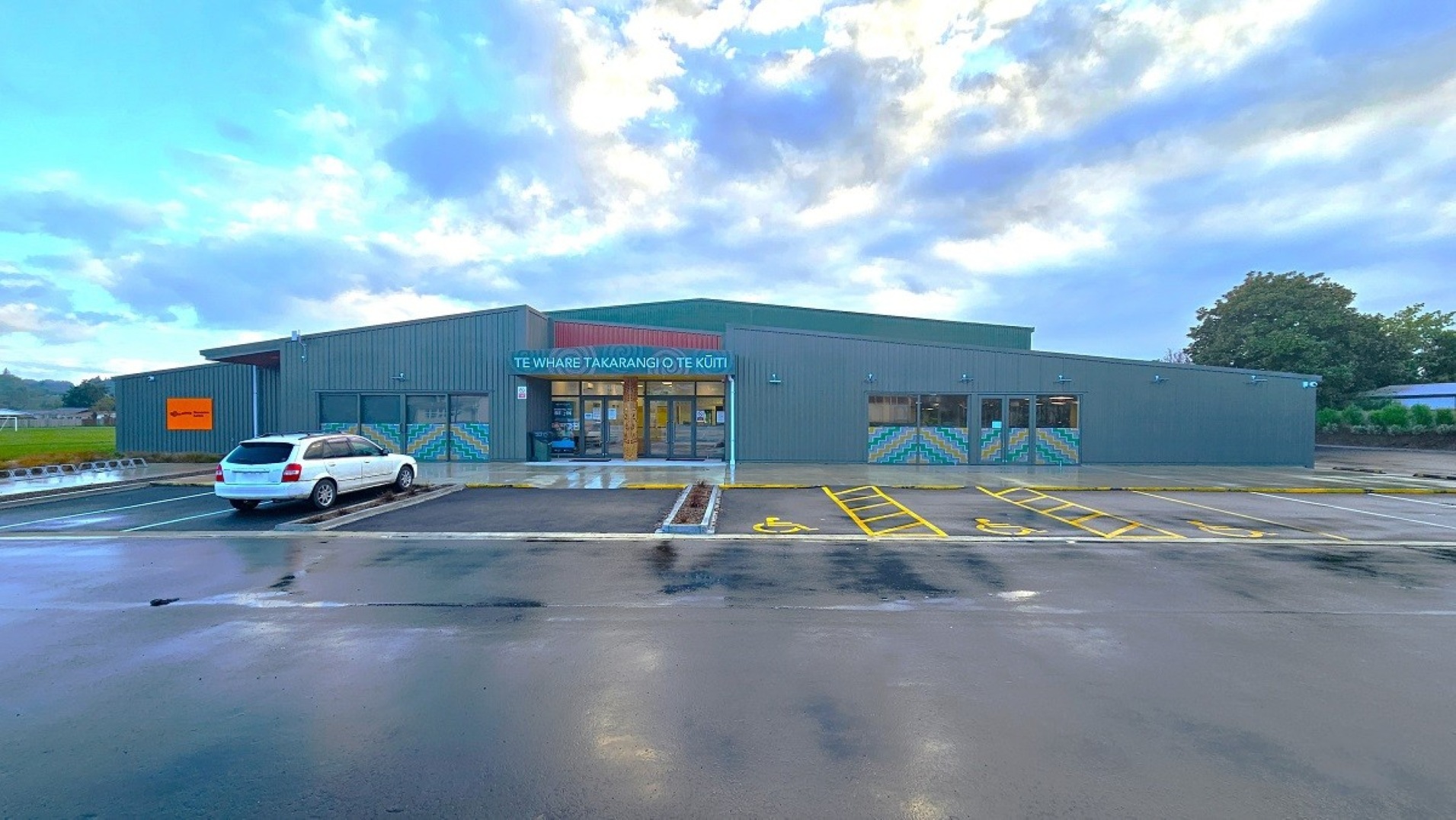 A modern, single-storey building with a grey exterior and a large entrance in the centre. The entrance has glass doors and a sign above it. The building is surrounded by a wet car park with marked parking spaces, including a white car parked on the left. The sky is overcast, and there are trees visible in the background on the right. The ground is wet, suggesting recent rain.