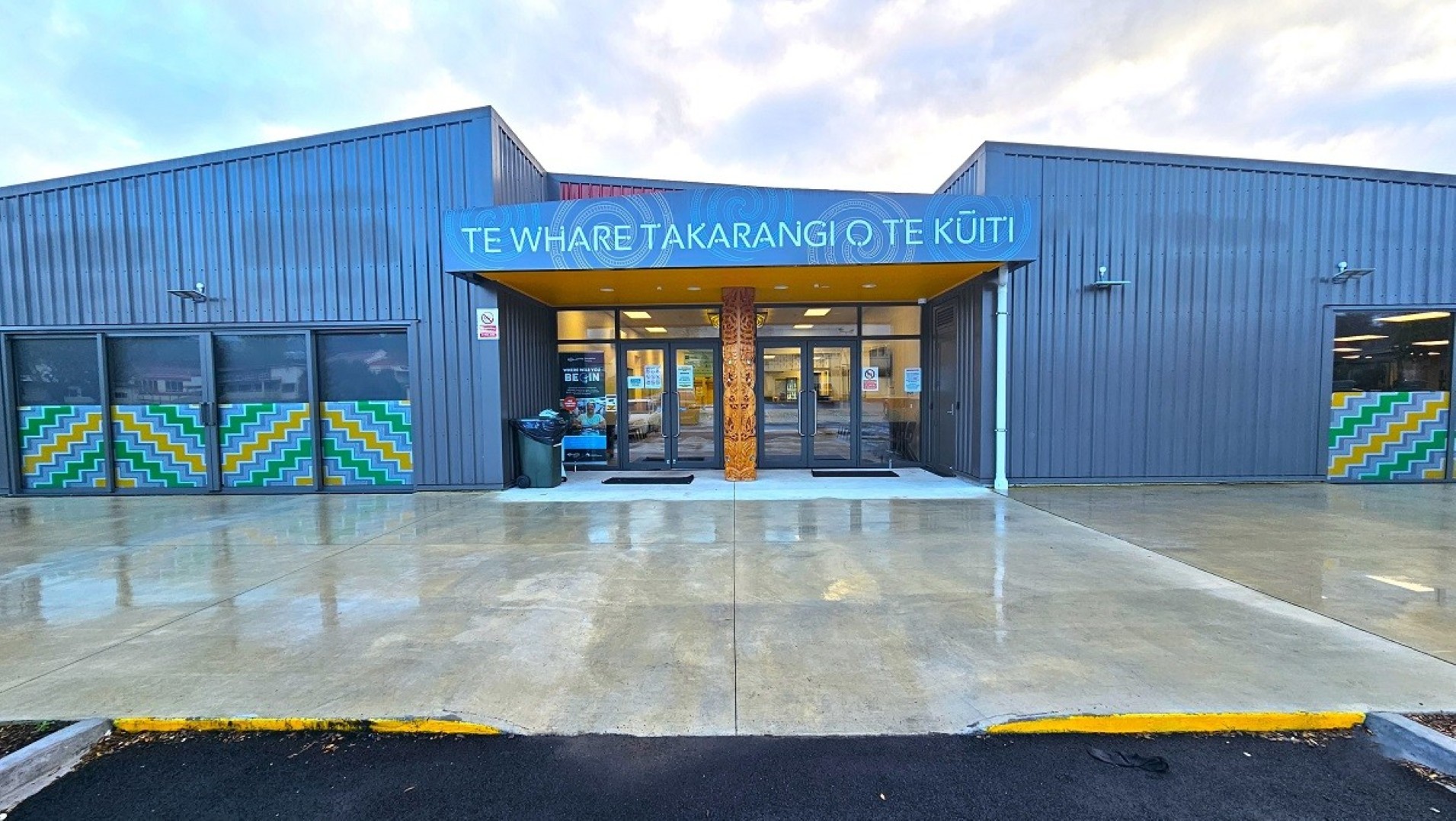 A modern building with a grey exterior and a blue sign above the entrance reading "Te Whare Takarangi o Te Kuiti". The entrance features large glass doors and a wooden carved pillar. On either side of the entrance, there are windows with colourful geometric patterns. The ground is wet, suggesting recent rain, and the pavement is smooth and reflective. The roof is flat, and the overall structure is symmetrical.