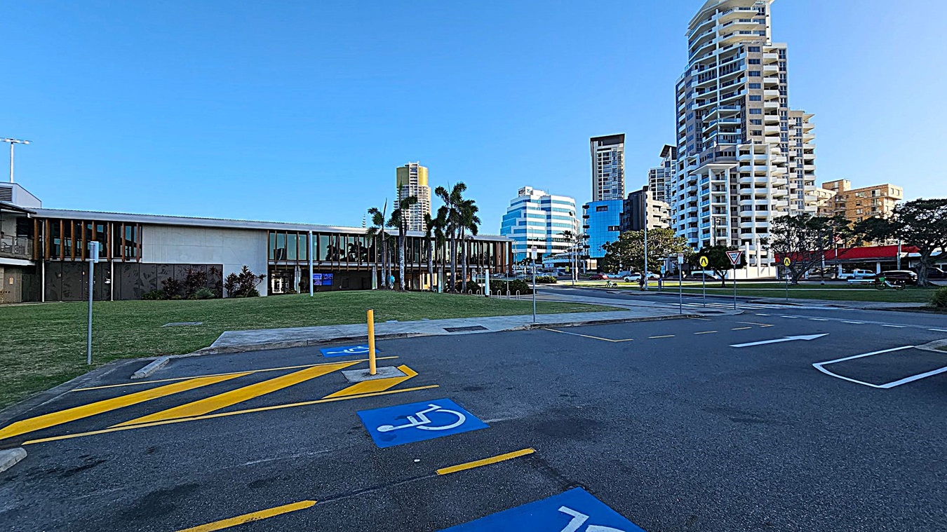 A parking area with marked disabled spaces in the foreground. On the left, there's a low, modern building with large windows and palm trees in front. In the background, several tall buildings with varied architectural styles rise against a clear sky. The ground is a mix of asphalt and grass, with a few road signs visible near the buildings.