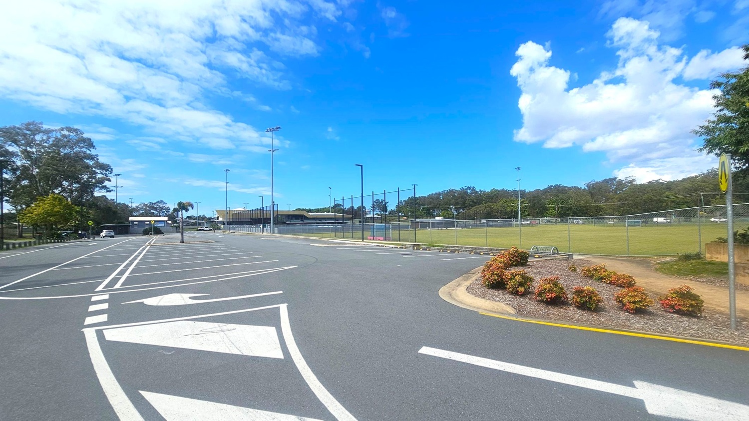 A car park with marked spaces and directional arrows on the asphalt. On the left, there are trees and a few parked cars. In the centre, a tall light pole stands near a fenced sports field. On the right, a landscaped area with small bushes and a signpost. The background features more trees and a building with a low roof.