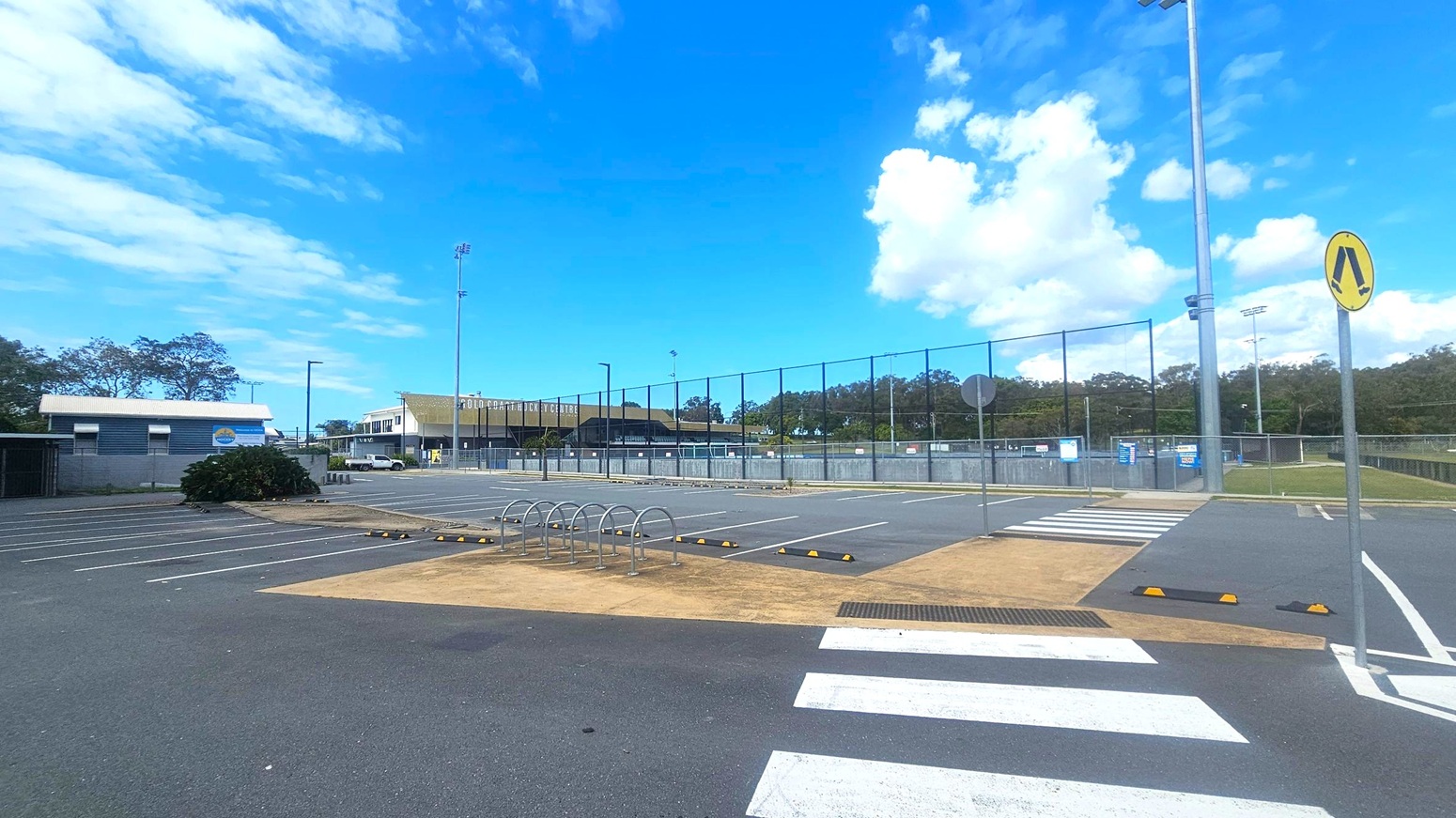 A car park with marked spaces and a bike rack in the foreground. A pedestrian crossing with a yellow sign is visible. In the background, there is a fenced sports field and a building with the words "Gold Coast Hockey Centre" on it. Tall floodlights are positioned around the area. Trees line the horizon, and the sky is clear with some clouds.