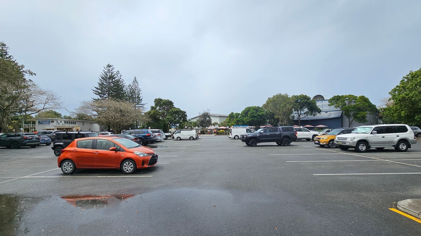 A car park with several vehicles, including an orange car prominently in the foreground. The ground is wet with a visible puddle reflecting the orange car. In the background, there are trees and a building with a sign partially visible. The area is surrounded by greenery, and the sky is overcast.