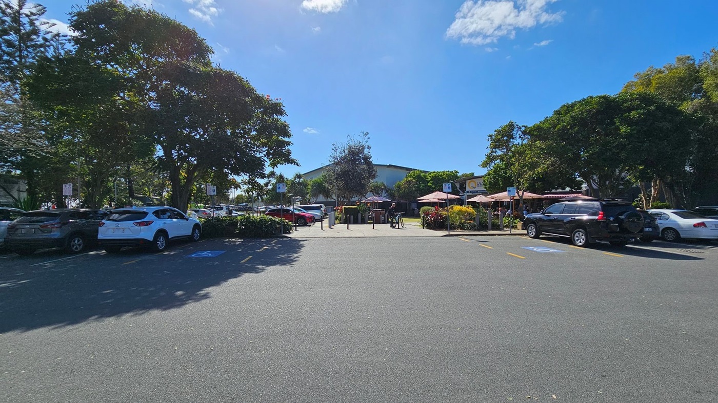 A car park with several vehicles, including a white SUV and a black SUV, is surrounded by trees and greenery. In the background, there are outdoor seating areas with umbrellas and a building with a sign that reads "QUE." A person is standing near a bicycle. The ground is paved, and there are marked parking spaces, including a disabled parking spot.
