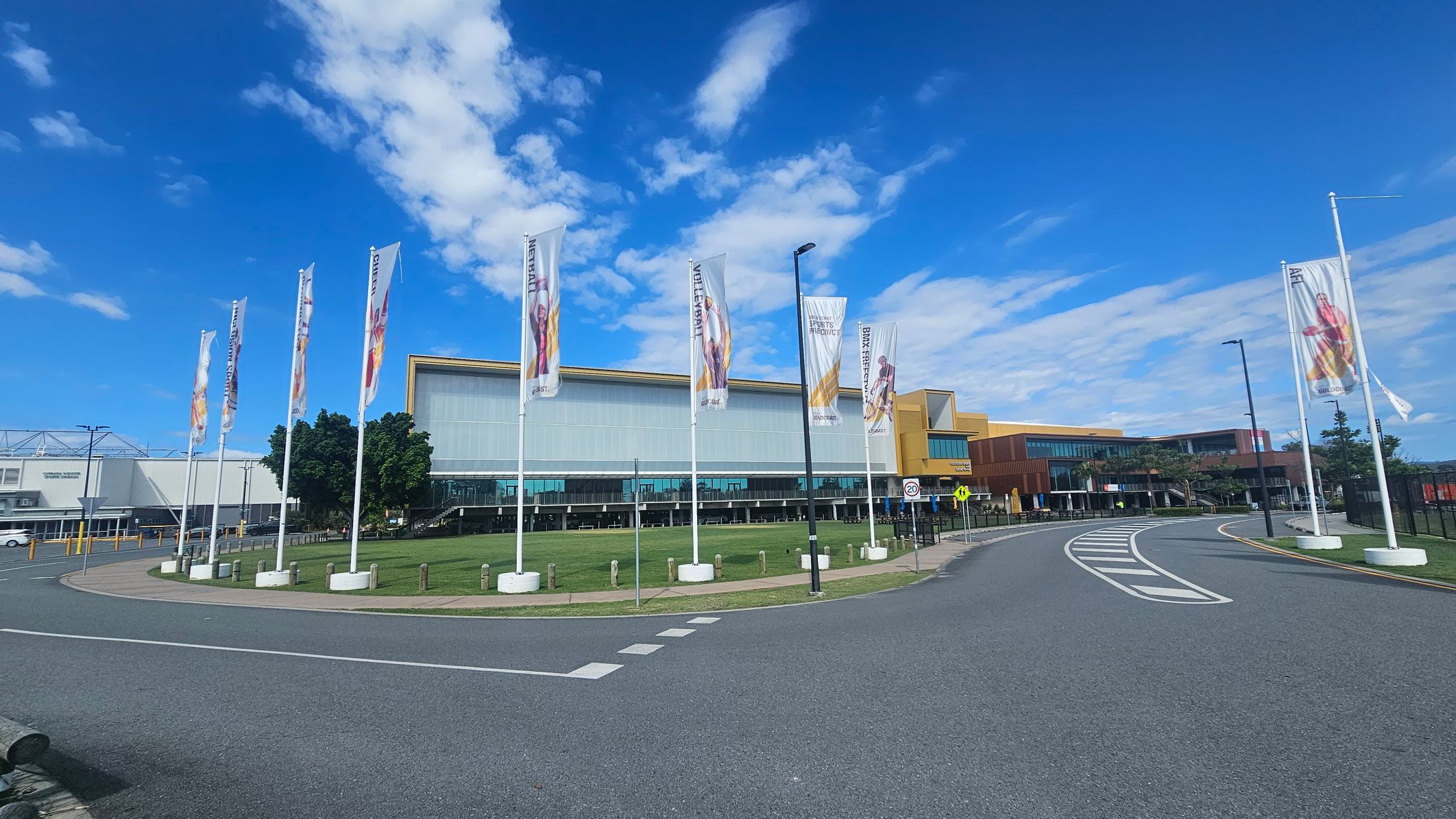 A modern building with a large glass facade is in the centre, surrounded by a series of tall flags on white poles. The flags display various images and text. To the left, there is a smaller structure with a grey roof. A curved road with white markings runs in front of the building, bordered by a grassy area with trees. The sky is clear with some clouds.