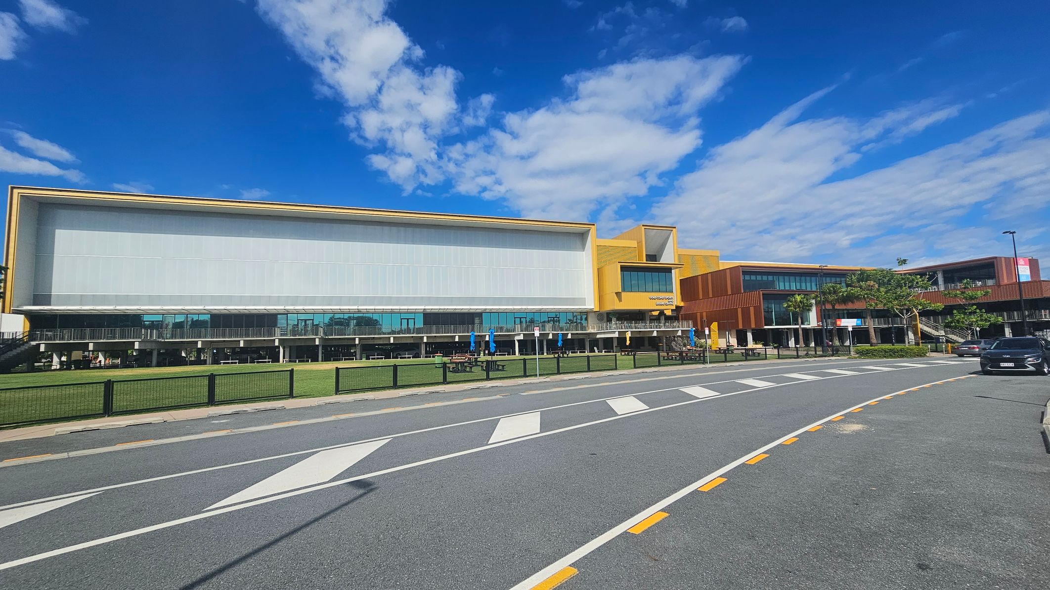 A modern building with a large glass facade and yellow accents on the right. The structure is two-storeyed with a mix of glass and wooden panels. In front, there's a grassy area with picnic tables and umbrellas. A road with marked pedestrian crossings runs parallel to the building, and a black car is parked on the right side. The sky is clear with some clouds.