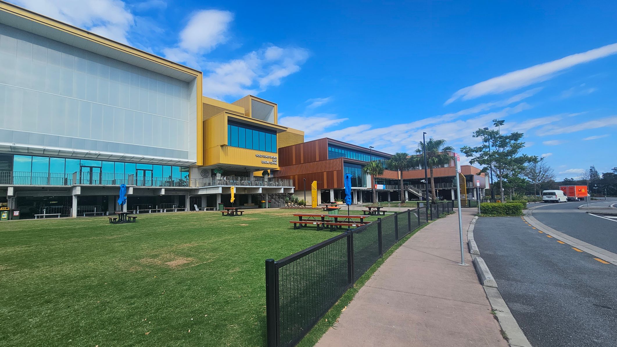 A modern building with a mix of yellow and brown panels on the left, featuring large glass windows. In front, there's a grassy area with picnic tables and blue umbrellas. A black metal fence separates the grass from a paved footpath on the right. Further right, a road curves with a white car and a red vehicle parked near some trees and shrubs.