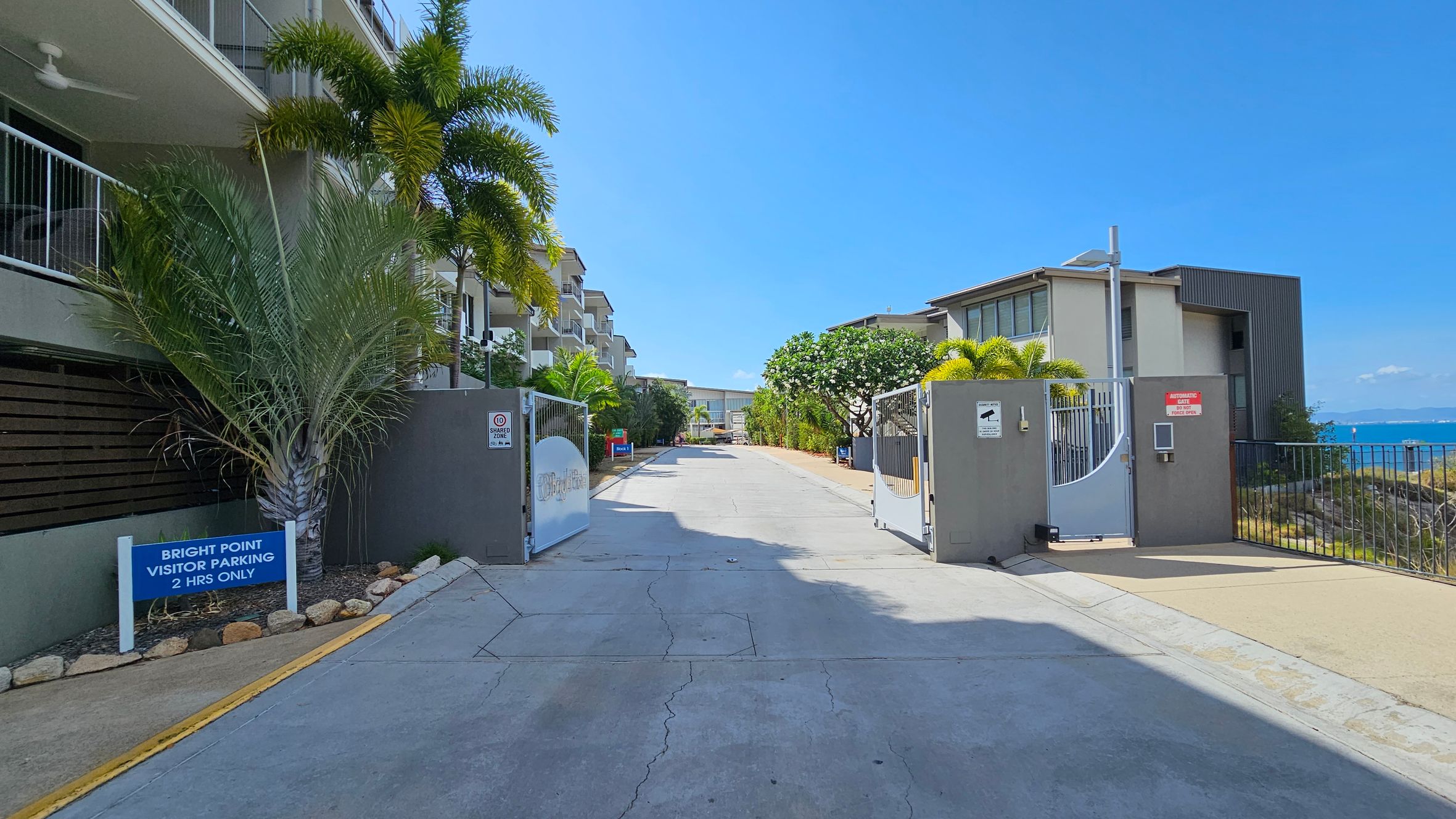 A gated entrance to a residential complex with a sign on the left reading "Bright Point Visitor Parking 2 Hrs Only." The left side features a building with balconies and palm trees. On the right, there's another building with a view of the ocean in the background. The ground is paved, and the gates are open, allowing a view down the driveway lined with more greenery and buildings.