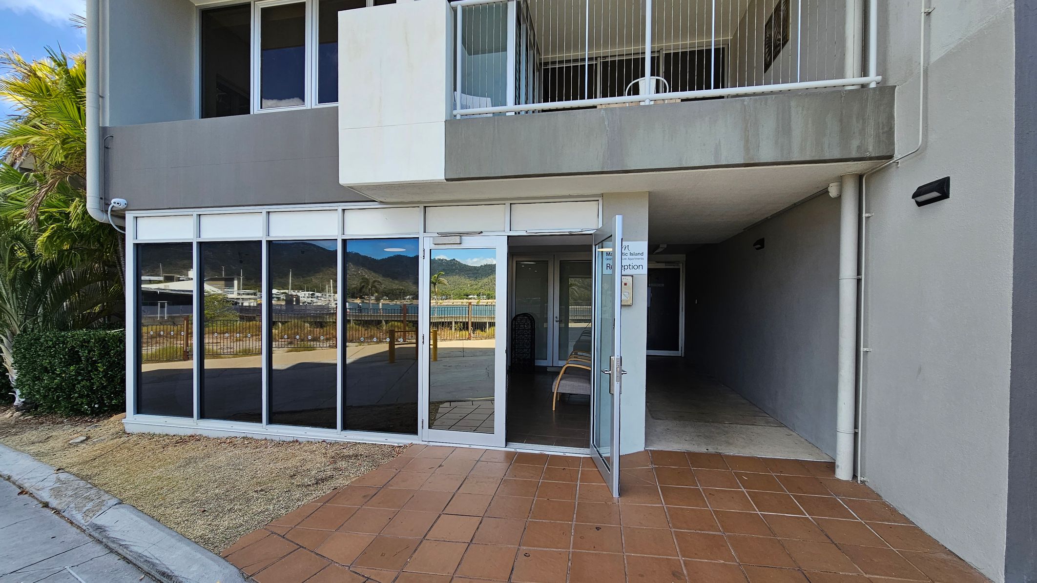 A building entrance with large glass windows on the left, reflecting a view of mountains and a marina. The entrance door is open, leading into a shaded interior space. Above the door, there's a balcony with a white railing. The ground is covered with terracotta tiles, and there's a small patch of grass and a bush to the left. A sign near the door reads "Reception". The building exterior is grey with a simple, modern design.