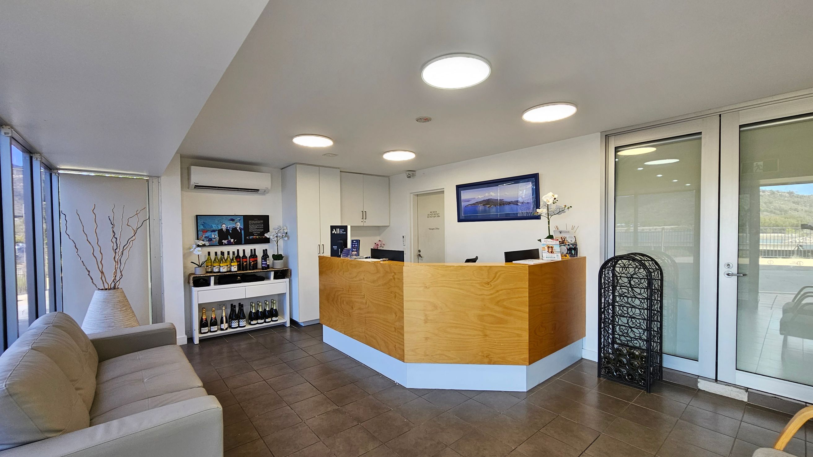 A modern reception area with a wooden desk in the centre, featuring two computer monitors. Behind the desk, a white wall has a framed landscape picture. To the left, a beige sofa sits near large windows, and a shelf displays various wine bottles. An air conditioning unit is mounted above the shelf. On the right, glass doors lead outside, with a decorative black screen nearby. The floor is tiled, and the ceiling has circular lights.