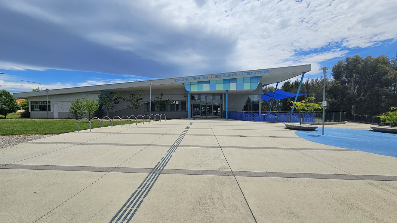 The building is a low-rise concrete structure with contrasting blue trim, and the facility name is displayed at the entrance. The building's concourse features a spacious concrete area. On the left, bike racks are positioned near the grass area. To the right, two small trees are in large concrete pots. The concrete floor has blue markings. The building has a blue fence railing and blue shade cloths. Tall trees fill the area on the right.