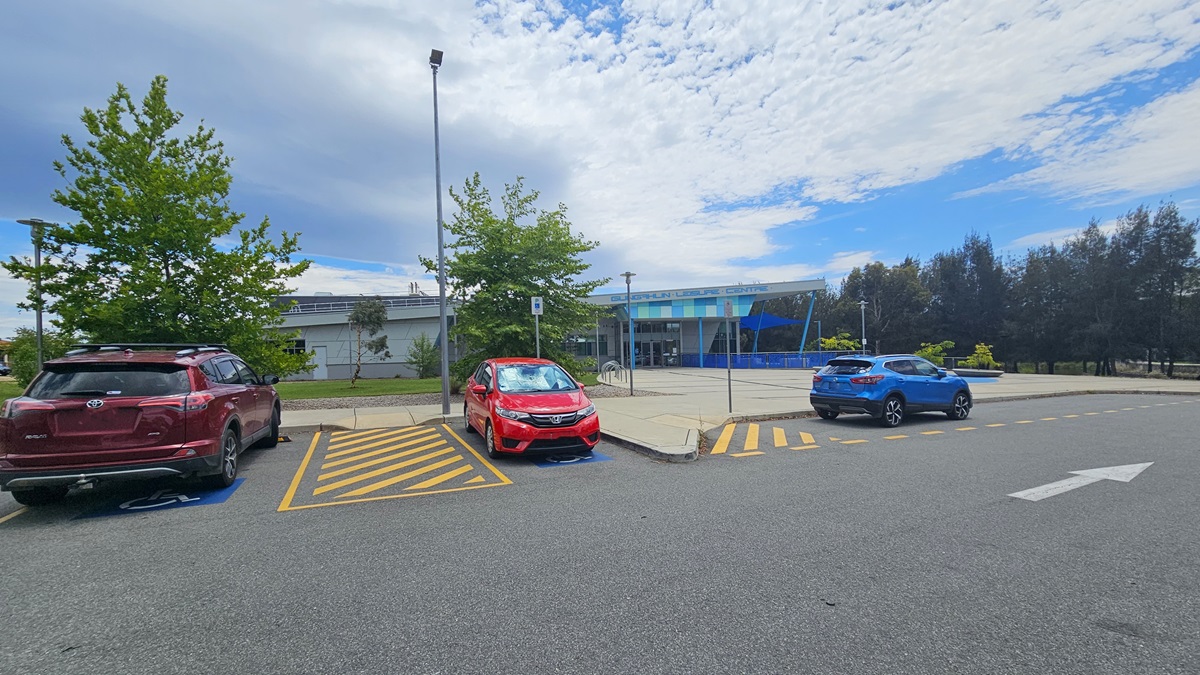 The car park features accessible parking slots marked with blue wheelchairs and yellow diagonal lines. Several cars of various models and colors are parked. On the right, a step-free ramp leads to the spacious building concourse. The building is a low-rise concrete structure with contrasting blue trim. Tall trees surround the area.