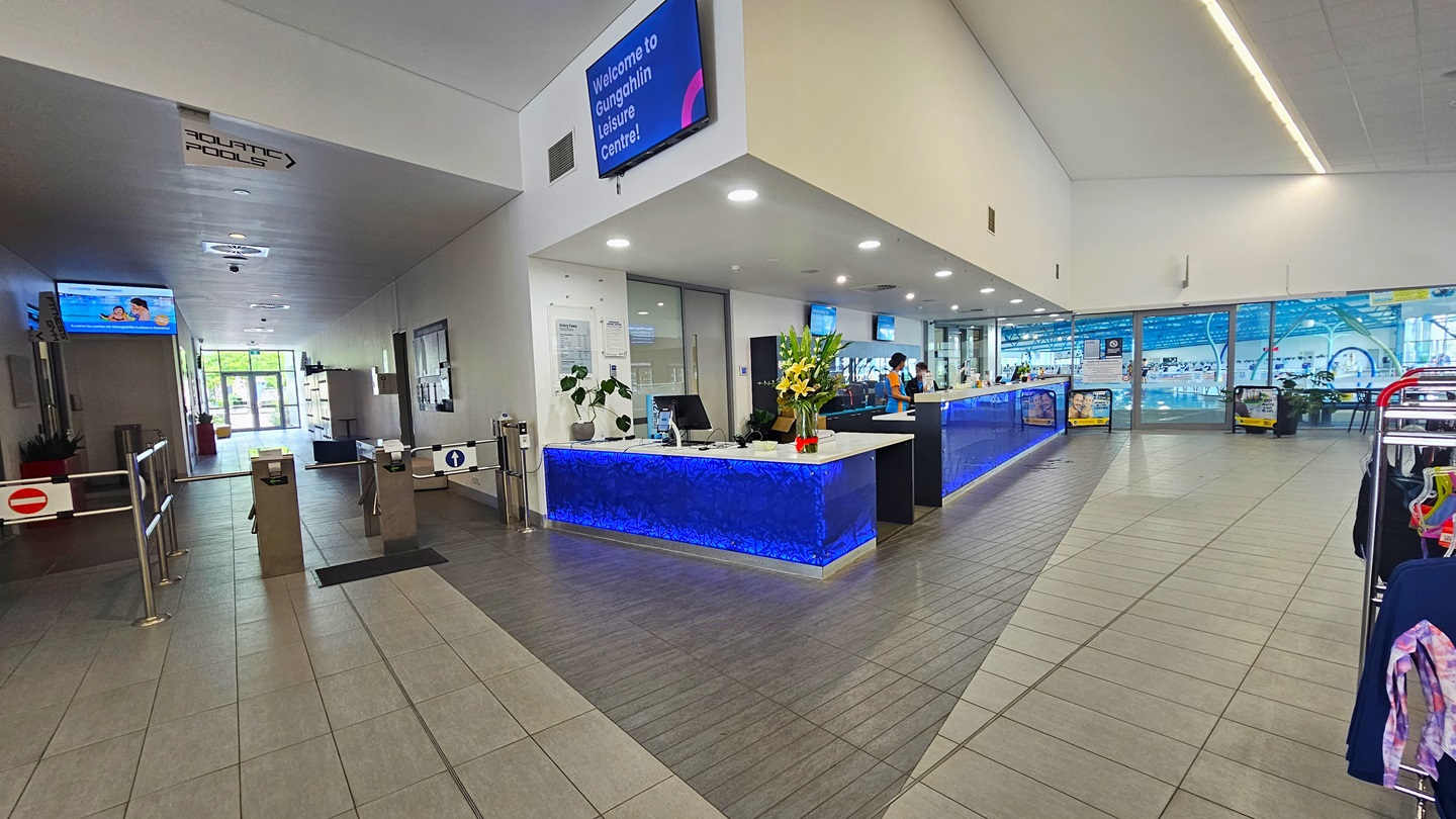 In the middle is the reception desk, featuring distinctive royal blue panels, adorned with flowers and indoor plants, with staff behind the counter. On the right, a retail area displays swimwear and accessories. To the left is a turnstile entry leading to the locker area. Flatscreen TVs are mounted on the wall. To the far left is the indoor aquatic facility. The ceiling is all white with fluorescent lights, and the floor has grey tiles.