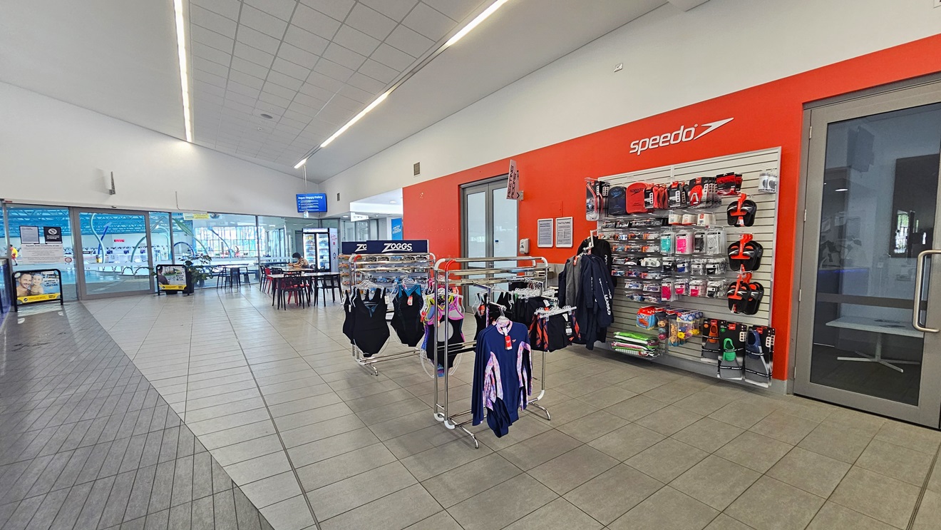 To the right is a retail area featuring swimwear, goggles, slippers, and other swimming accessories. A manual glass door is positioned next to it. Movable tables and chairs are arranged on the right. A fridge with cold drinks is positioned in the corner, and on top of it is a flatscreen TV. The indoor pool is ahead, and banners are visible near the entrance. The floor has grey tiles, and the white ceiling has a design resembling the floor tiles below it.