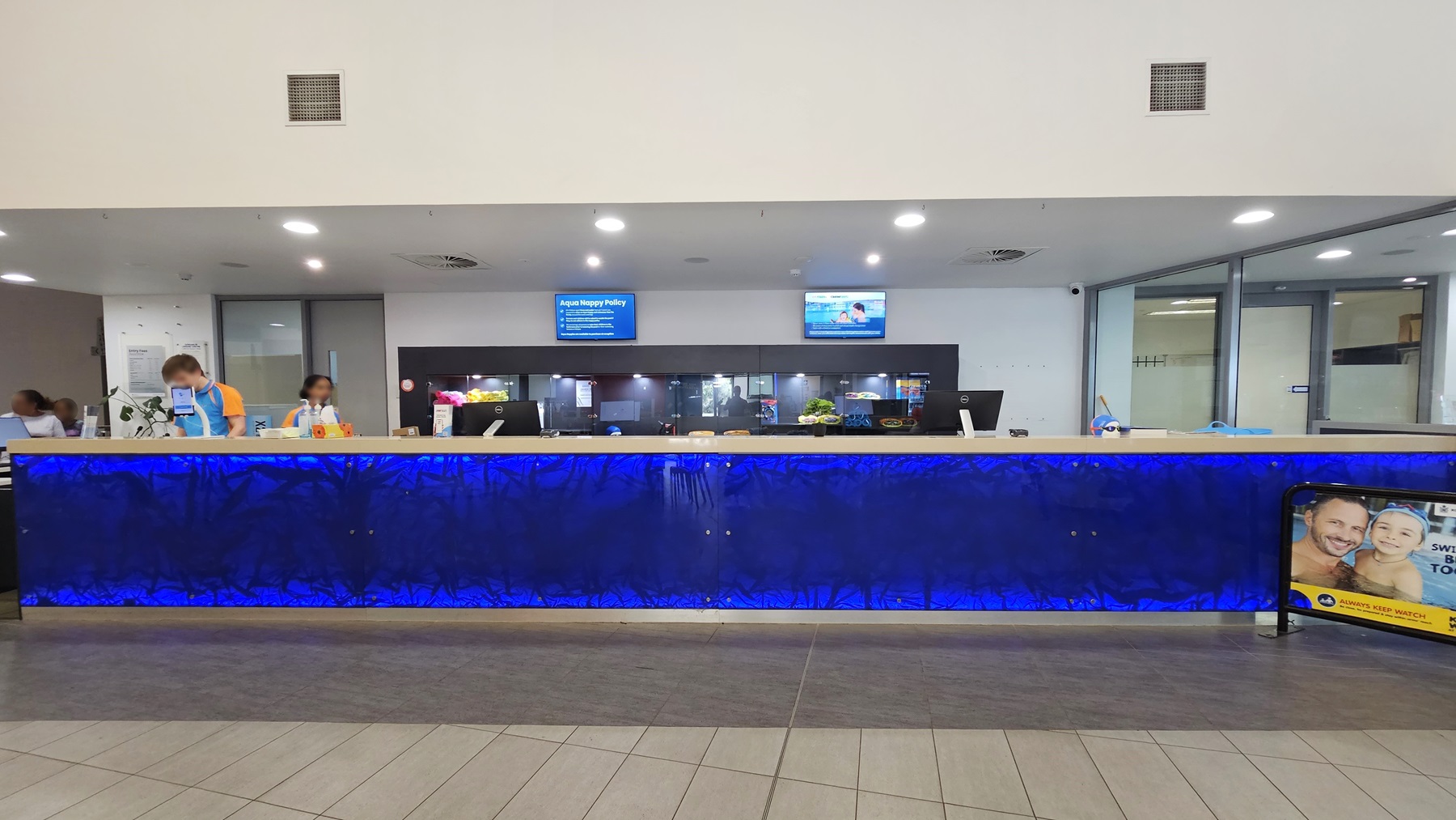 In the middle is the reception desk, featuring distinctive royal blue panels with staff behind the counter. A display rack is positioned behind the counter. Two flat screen TVs are mounted on the wall. A banner is positioned on the right. The ceiling is all white with fluorescent lights, and the floor has grey tiles.