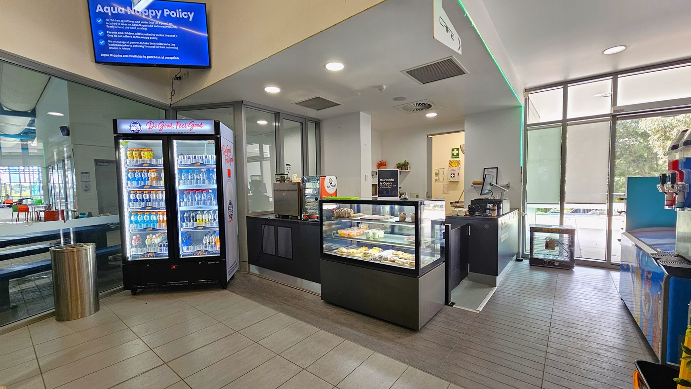 In the middle is a cafe featuring a food rack with fresh salad bowls, sandwiches, and wraps. To the left is a fridge filled with cold drinks. A silver rubbish bin is next to it. To the right is an ice cream freezer and a beverage dispenser. The glass wall on the right allows a view of the outside. Movable chairs and tables in bright shades of green and orange are arranged. A flat-screen TV is mounted on the middle wall near the white ceiling.