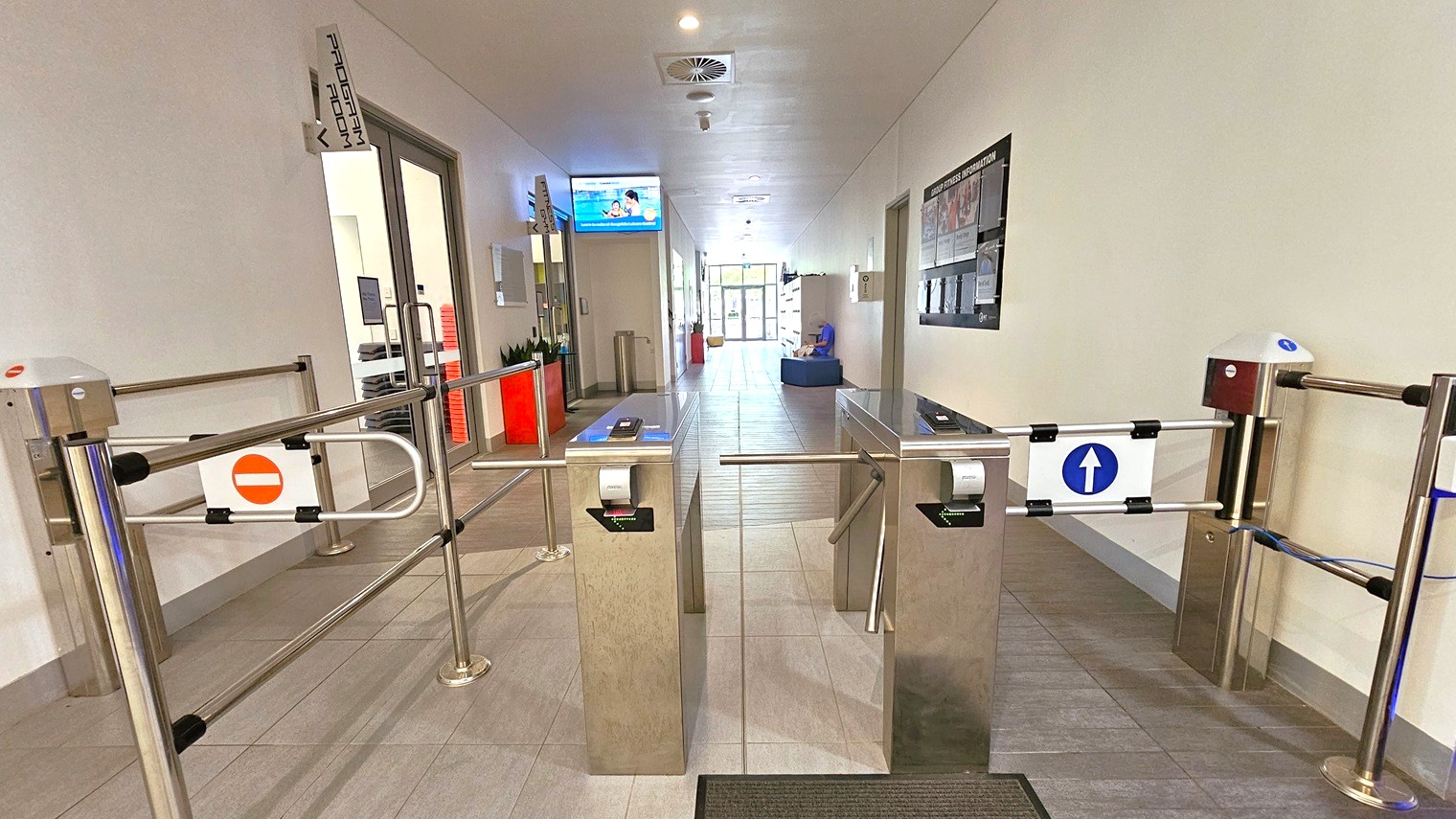 Two turnstiles are positioned at the entrance—one on the left for exit and the other on the right for entry, each with clear markings. Upon entry, the Program Room is on the left, and to the right is a blackboard with posters. A flat-screen TV is mounted on the left wall near the ceiling, above a plant box and a rubbish bin. To the far right is the locker area, cushioned seating, and a glass door. The ceiling and walls are white, and the floor features grey tiles.
