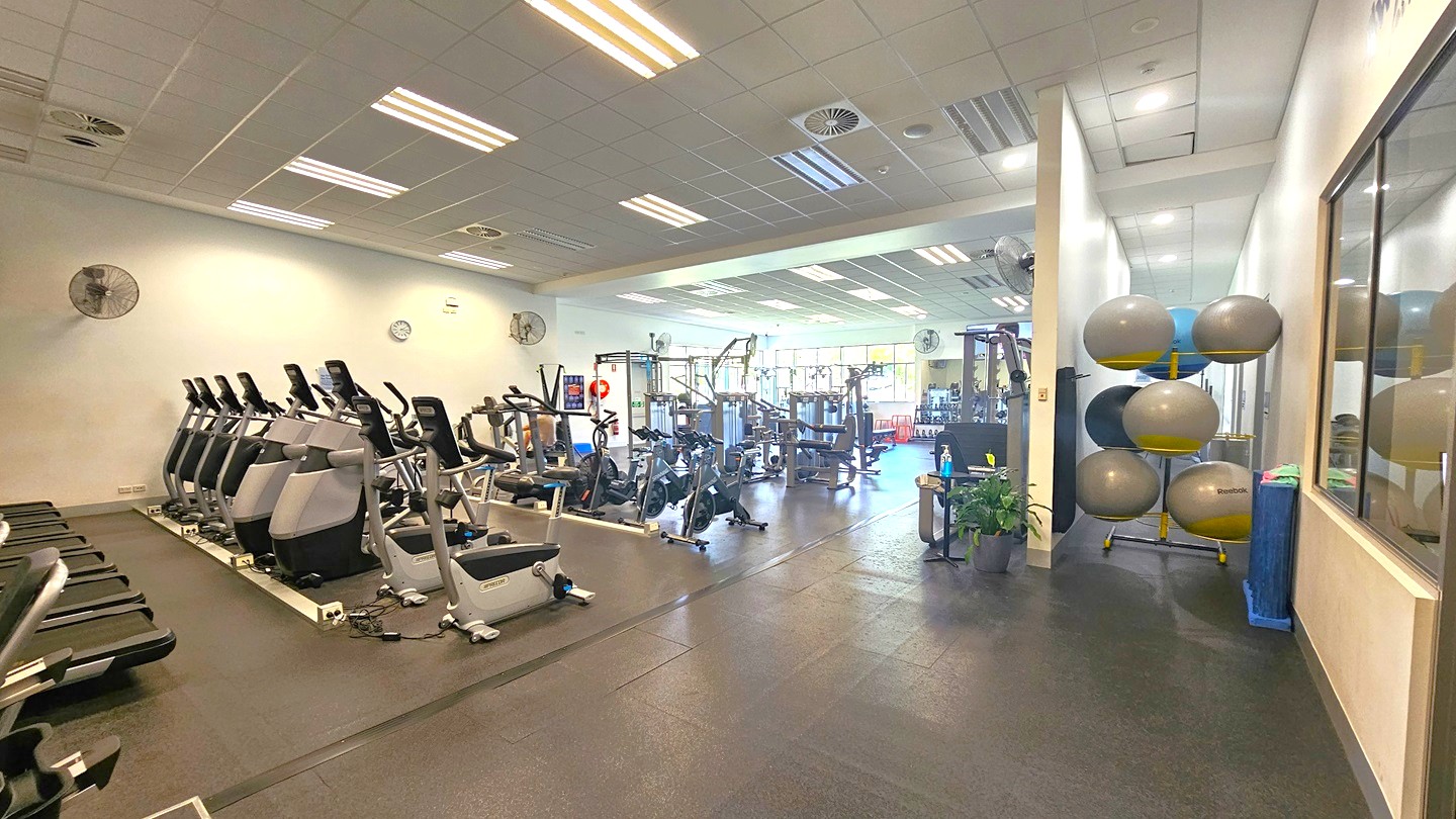 Inside the gym, cardio and strength equipment lines the left side—smith machines, Lat Pulldown machines, and more. Neat stacks of stability balls fill the right. A dividing wall separates areas. Flat-screen TVs and wall fans are fixed to the walls. The ceiling exhibits a white porous design, lit by fluorescent lights. The floor features black rubber tiles. On the right, a window with a glass wall provides natural light.