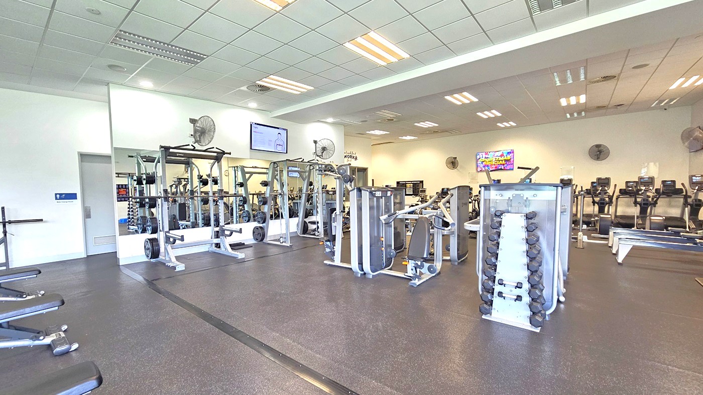 On the left, are bench press machines; in the middle, Smith Machines by a mirror, surrounded by free weights and resistance machines. On the right are row machines and cardio machines. An accessible bathroom is located on the left, behind the middle wall. Black rubber tiles cover the floor. Flat-screen TVs and wall fans are mounted on the upper wall. The ceiling is uniformly white, adorned with fluorescent lights.