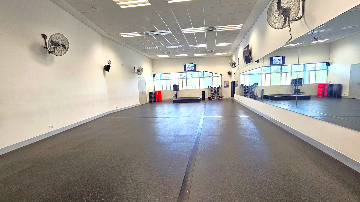 A spacious gym room with a dark rubber floor and a high ceiling with fluorescent lights. The left wall has several mounted fans and speakers. The right wall is covered with large mirrors reflecting the room. At the far end, there are large windows allowing natural light in, and a small platform with exercise equipment like weights and mats. A television is mounted above the windows.