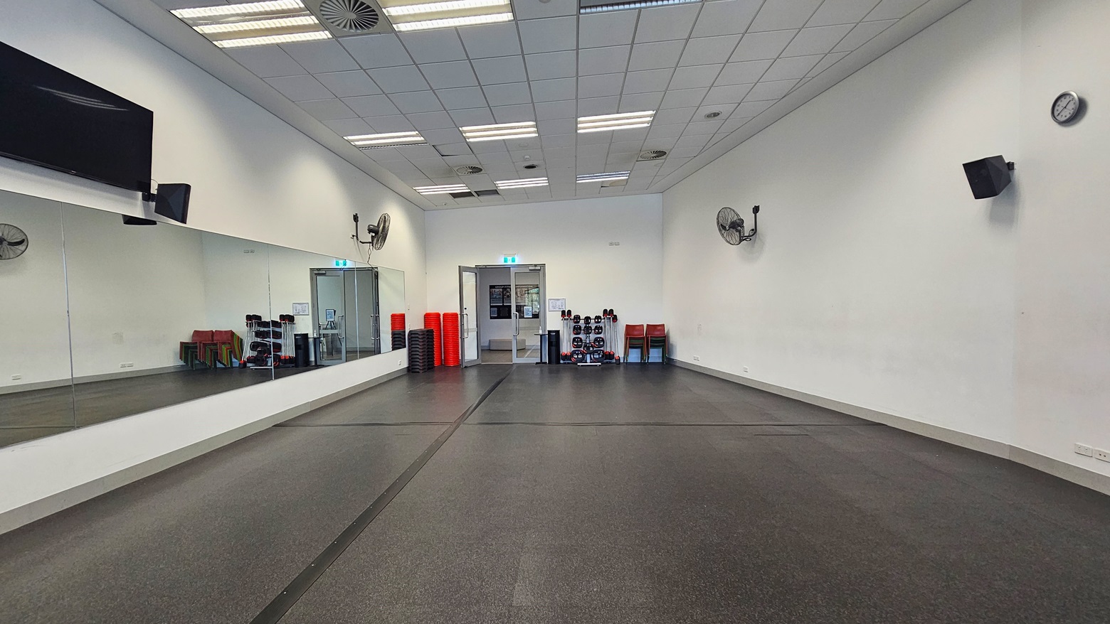 The fitness room is spacious, with black rubber tiles covering the floor. Mirrors line the left wall, while a flat-screen TV, wall fans, a clock, and audio speakers are affixed to the upper walls. In the middle, an inward-opening glass door is present. Fitness equipment is arranged near the door, alongside red plastic chairs. The ceiling and walls are consistently white.