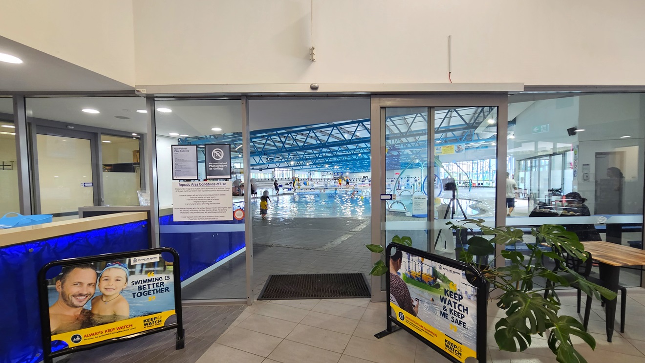In the middle is an automatic glass sliding door. Posters are affixed to the left door. A section of the blue reception desk is visible on the left. Banners are positioned on both sides. Movable tables and chairs, along with indoor plants, are positioned on the right. Inside, the Kids Pool is visible. Exposed blue beams dominate the ceiling. Swimmers are using the pool.