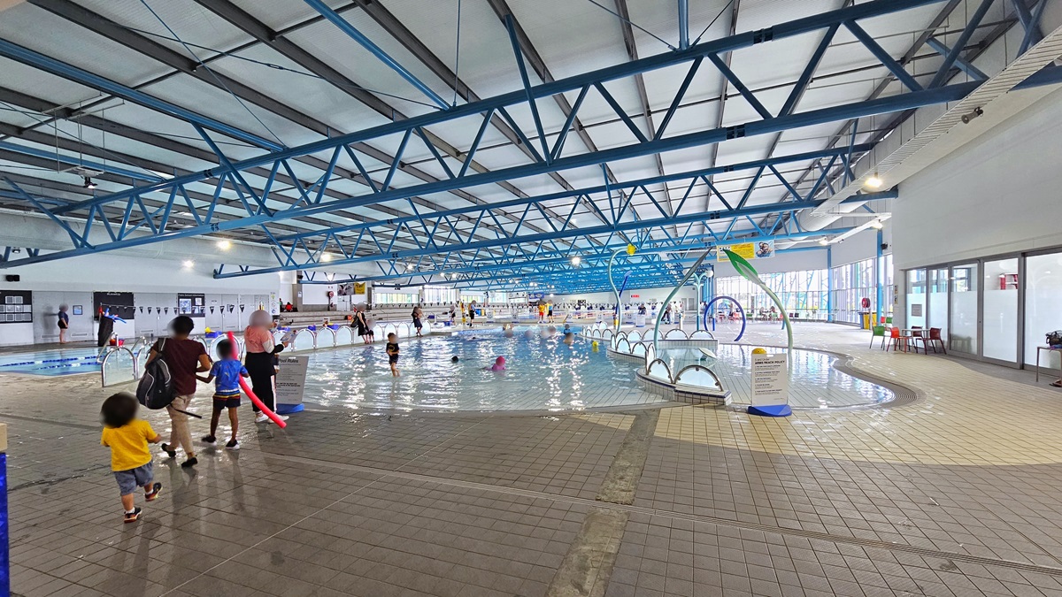 In the middle is the Toddler Splash Pool, equipped with water sprays and fountains. Adjacent to it is the Training Pool, separated by half-circle fence railings. To the left is the lap pool, equipped with floating ropes. On the right, movable plastic chairs and tables are positioned near the glass wall. The ceiling features a white industrial look with exposed blue metal beams. The pool is occupied by guests.