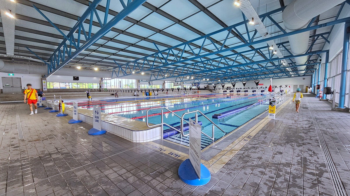 The 50m Lap pool features stairs and ramps with handrails on each side, along with floating ropes. To the left, free-standing banners line the pool deck, and tiered seating is available on the far left. Glass walls surround the area, and the ceiling has an industrial look with exposed black and blue metal beams, large aircon vents, and LED lights. Two people are seen on the pool deck.