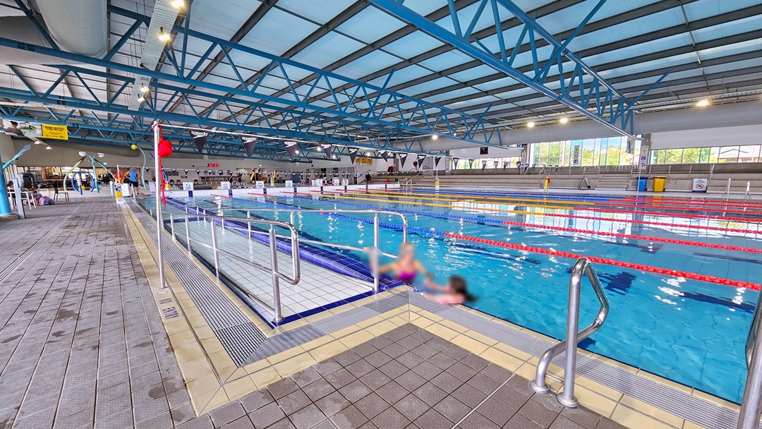 The 50m Lap pool has a ramp entry with handrails and floating ropes. To the far left is the kids' splash pool. Free-standing banners line the pool deck, and tiered seating is available on the far right. Glass walls surround the area, and the ceiling has an industrial look with exposed black and blue metal beams, large aircon vents, and LED lights. Two people are seen on the pool deck. Poles hold a string banner that runs across the pool.