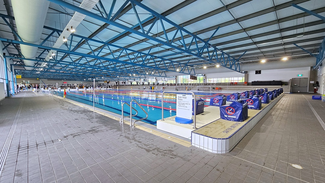 The 50m Lap pool features a ramp entry with handrails and floating ropes. To the right, diving boards with blue covers are arranged on the raised pool deck. A handrail is installed on the corner of the pool. To the far right is tiered seating, and to the far left is the kids' pool. Poles hold a string banner that runs across the pool. Glass walls surround the area, and the ceiling has an industrial look with exposed black and blue metal beams, large aircon vents, and LED lights.