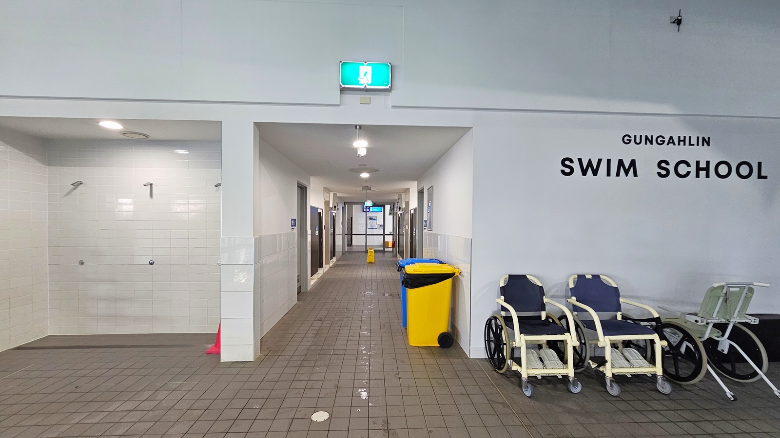 To the left are three showers. Directly ahead is a corridor leading to the bathroom's. A yellow and blue rubbish bin are on the right of the corridor. Three aquatic wheelchairs are placed to the right of the entrance to the corridor. 'Gungahlin Swim School' is written in black on the wall above the wheelchairs.