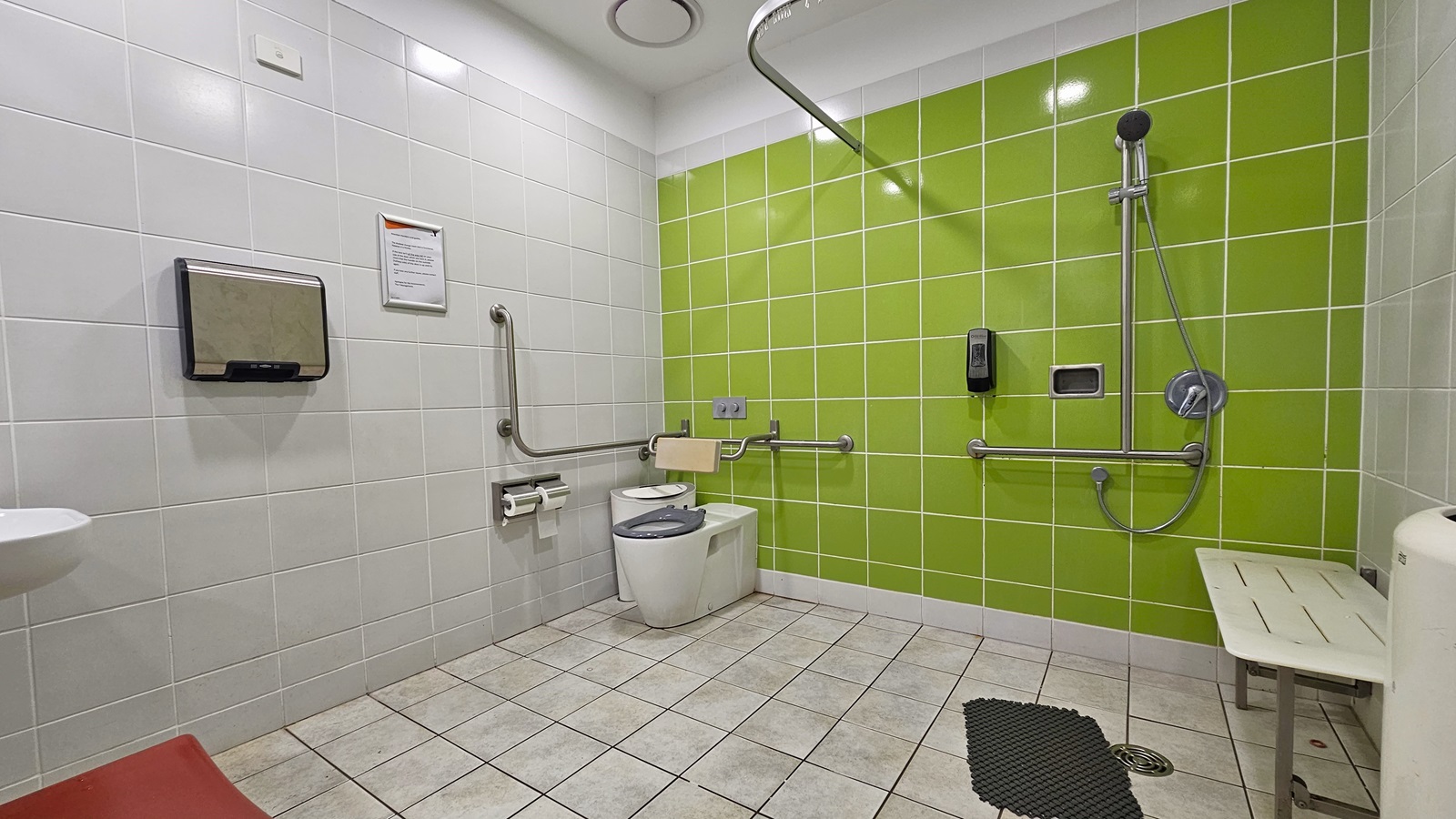 Inside an accessible bathroom with a vivid lime green tiled wall and a grey tiled wall. To the right is a bench seat adjacent to the shower. Directly ahead is the toilet and to the left is the hand dryer and a glimpse of the handbasin