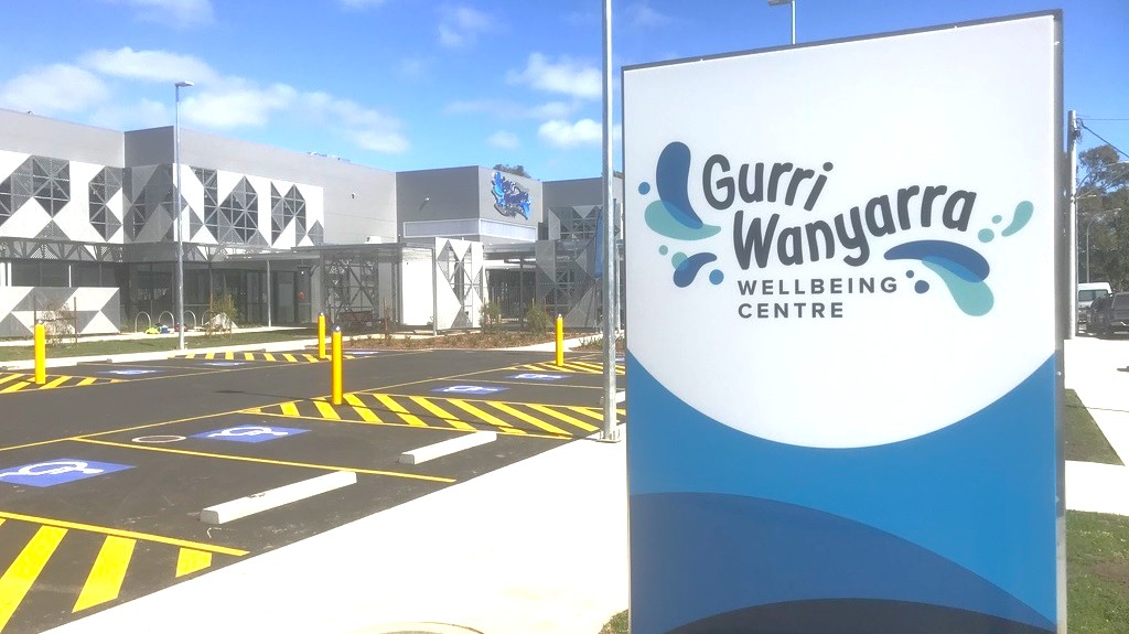 A large sign on the right reads "Gurri Wanyarra Wellbeing Centre" with blue and teal water droplet graphics. On the left, a modern building features geometric patterns on its facade. The foreground shows a car park with yellow-striped markings and several yellow bollards. The background includes a clear blue sky.