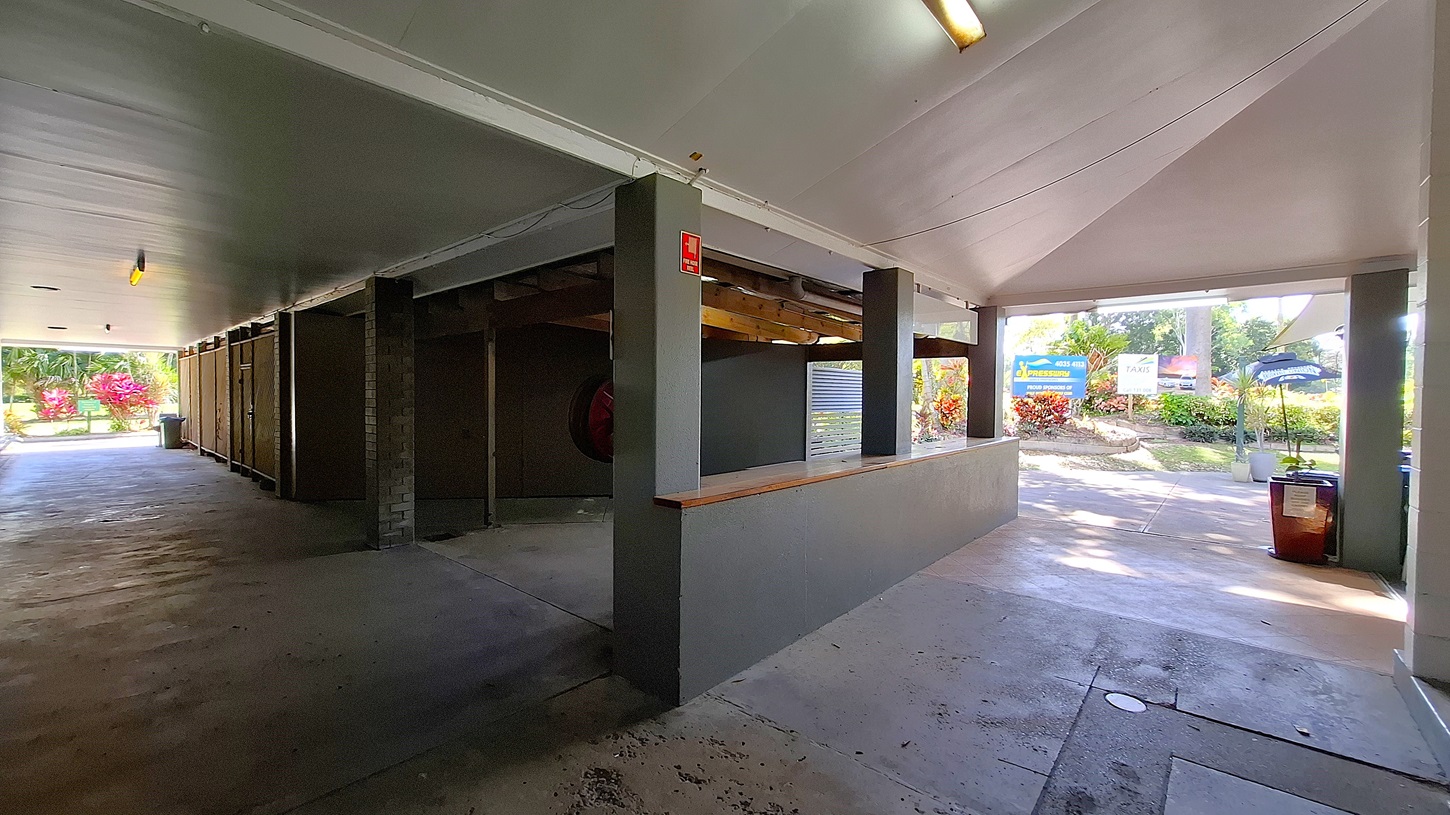 On the left, a concrete wall features a series of storage units, with a fire extinguisher mounted on the side. The floor is smooth and grey, extending towards an open area with a pathway leading outside. In the centre, a wooden counter runs along the wall, providing a surface for service or display. On the right, a large opening leads to a landscaped area with colourful plants and a sign visible in the background. The ceiling is flat and painted white, with fluorescent lights.