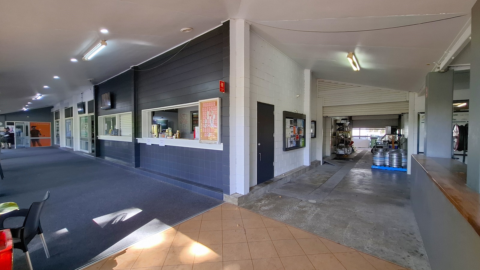 On the left, a long corridor features a dark carpeted floor, with several windows and a service counter displaying drinks and snacks. The walls are painted in light colours, with a few televisions mounted above. In the centre, the corridor leads to an open area with a tiled floor, transitioning to a concrete surface. On the right, a large opening reveals a storage area with kegs and equipment. The ceiling is flat and painted white, with fluorescent lights.
